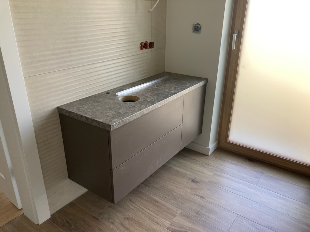 Szara szafka łazienkowa z blatem z otworem na umywalkę, zamontowana na ścianie z dekoracyjną fakturą, drewnopodobna podłoga, widok z boku, okno z matową szybą.