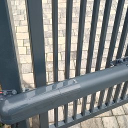 Usługi Ogólnobudowlane Stal-Pro - Szara brama metalowa z zamontowanym siłownikiem AVO, widok z bliska na mechanizm. W tle kostka brukowa i kwiaty w doniczce przy ogrodzeniu panelowym.