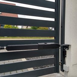 Usługi Ogólnobudowlane Stal-Pro - Szara, pozioma brama z automatyką, widoczny siłownik i skrzynka sterująca na tle jasnej ściany budynku. Widok z bliska na mechanizm bramy.