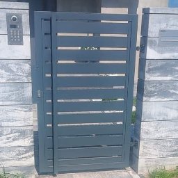 Usługi Ogólnobudowlane Stal-Pro - Nowoczesna, szara furtka z poziomymi szczeblami, wkomponowana w ogrodzenie z betonowych bloczków o marmurkowym wzorze. Widoczny domofon i skrzynka na listy.