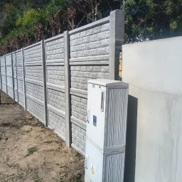 Usługi Ogólnobudowlane Stal-Pro - Długi, szary, betonowy płot z wzorem cegieł, oddzielający posesję od ulicy. Obok płotu skrzynka elektryczna i fragment białego muru. W tle drzewa i znak drogowy.