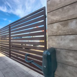 Usługi Ogólnobudowlane Stal-Pro - Nowoczesna, przesuwna brama z poziomych profili w kolorze brązowym, z automatyką AVO, zamontowana na słupkach z szarego betonu architektonicznego. Widok z zewnątrz.