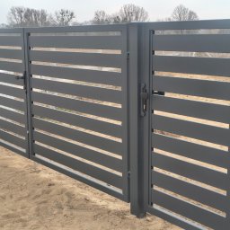 Usługi Ogólnobudowlane Stal-Pro - Nowoczesna, szara brama metalowa z poziomymi szczeblami, widoczny zamek. Tło stanowią drzewa bez liści i piaszczysta ziemia. Minimalistyczny design.
