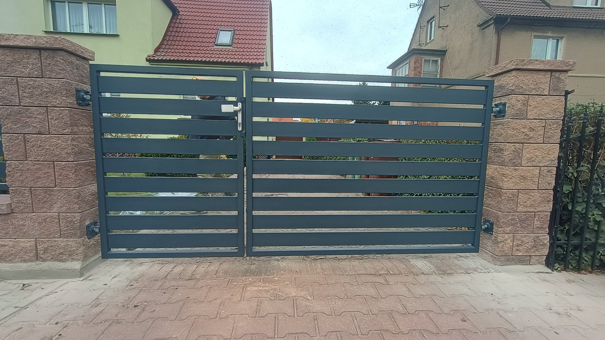 Nowoczesna, pozioma brama metalowa w kolorze grafitowym, zamontowana na murowanych słupkach z cegły, z widocznym zamkiem i klamką. Brama wkomponowana w otoczenie domu.