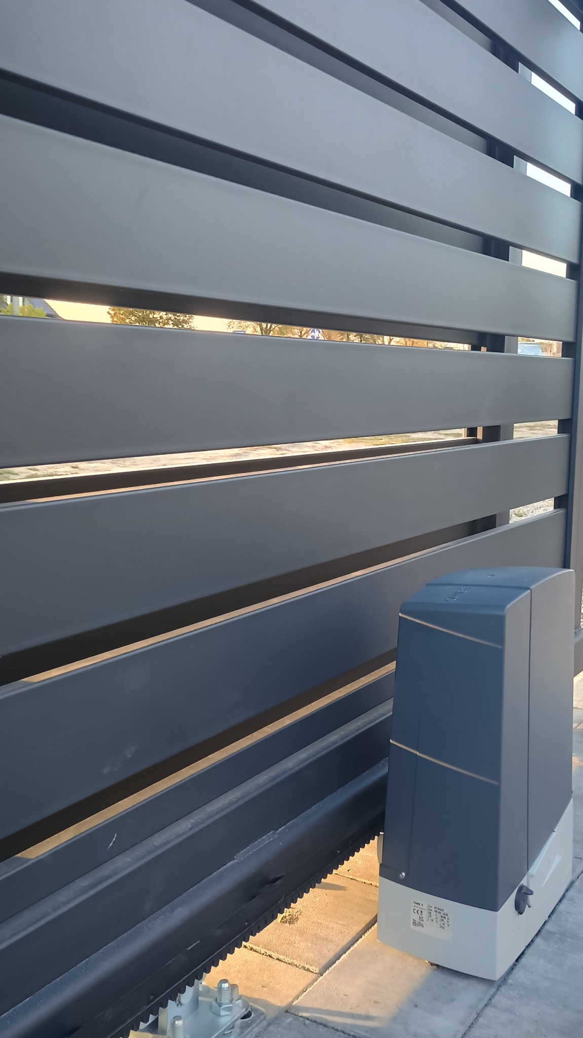 Szara, pozioma brama przesuwna z napędem elektrycznym CAME, widoczna z bliska, z zewnątrz. Widoczne elementy montażowe i zębatka.