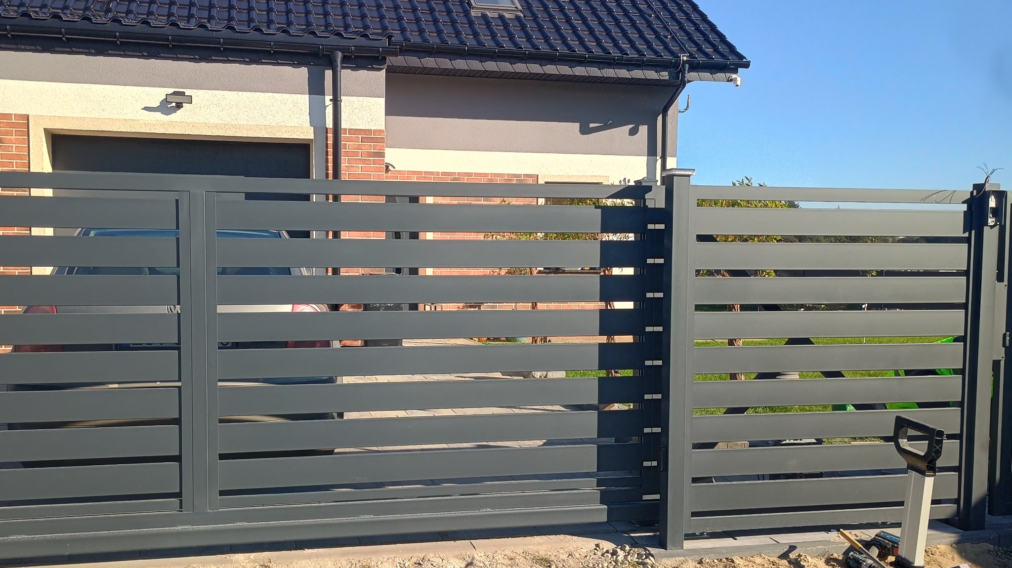 Nowoczesna, przesuwna brama w kolorze grafitowym z poziomymi panelami, na tle domu z ceglaną elewacją. Widoczne narzędzia sugerują trwający montaż.