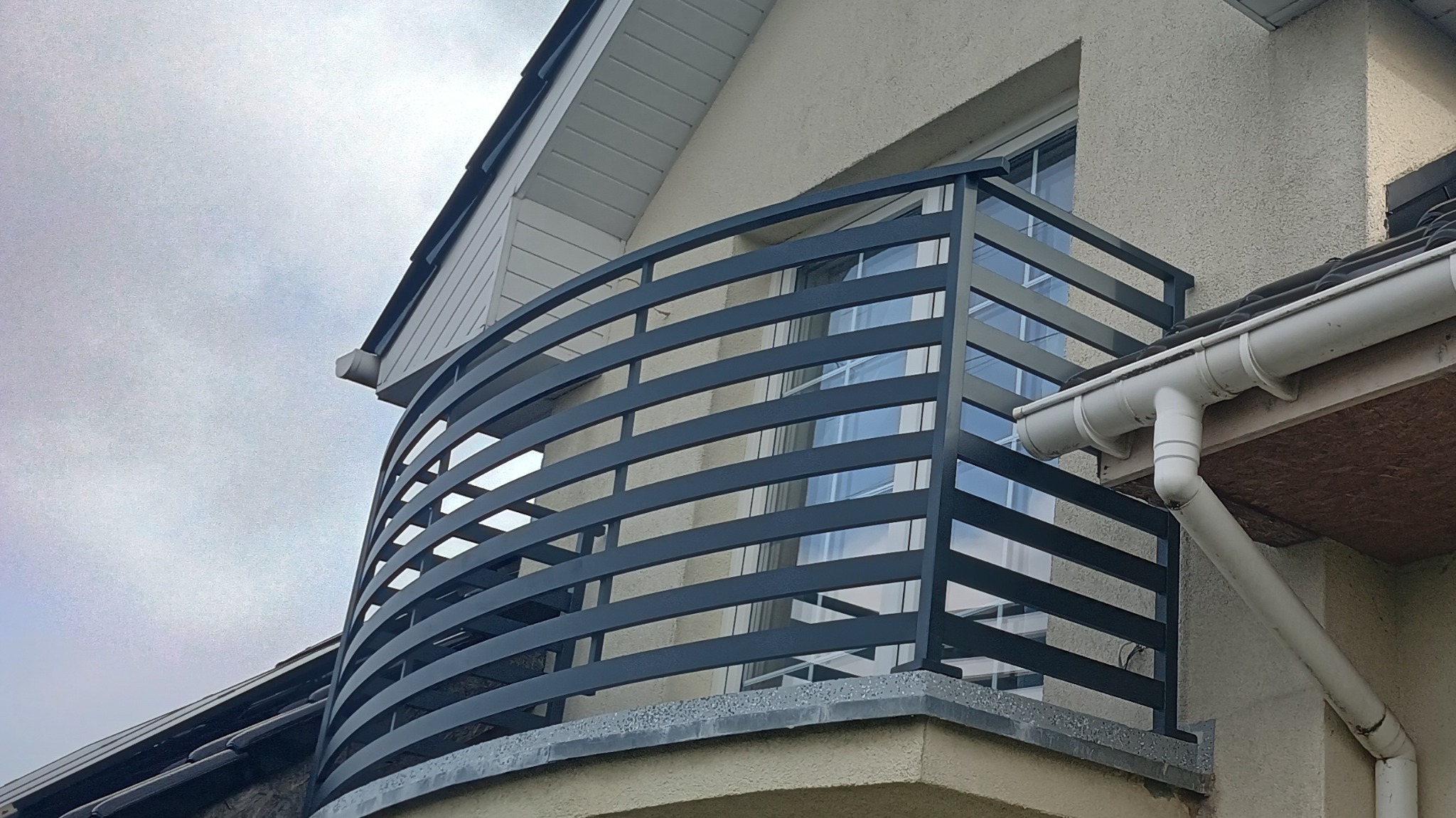 Nowoczesna, półokrągła poręcz balkonowa z metalowych, poziomych prętów, zamontowana w Stargardzie. Widok z dołu, na tle okna i fragmentu elewacji.