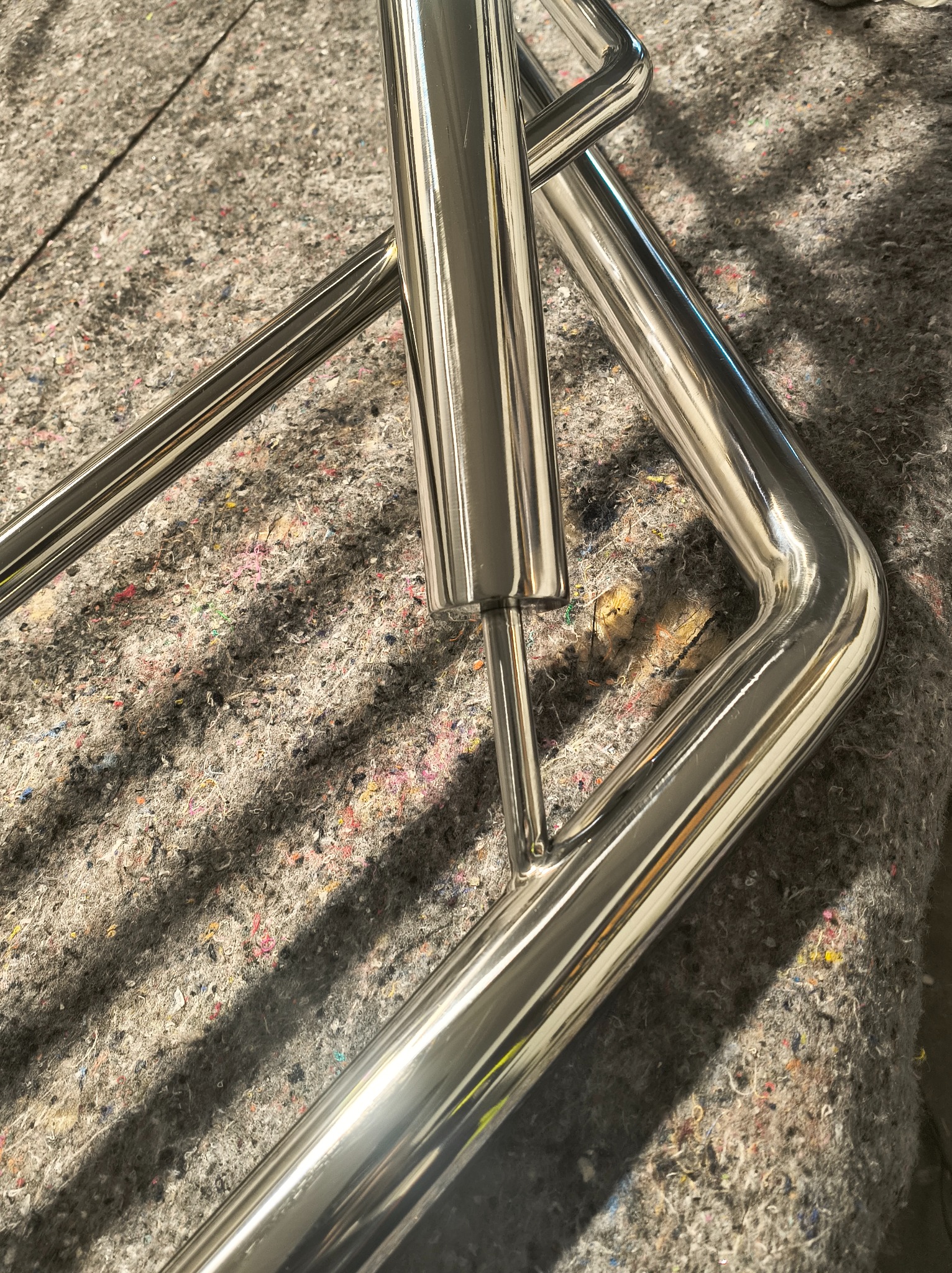 Element poręczy ze stali nierdzewnej, z widocznymi spawami, leżący na szorstkiej powierzchni. Metal polerowany na wysoki połysk, odbija światło.