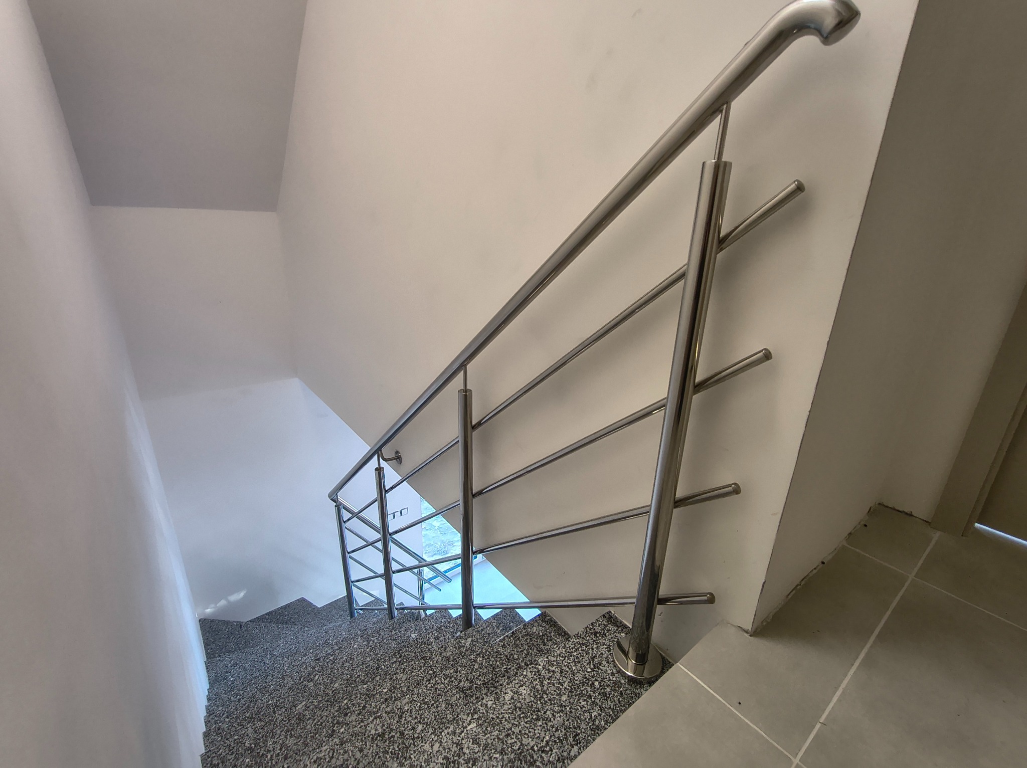Nowoczesna, metalowa poręcz schodowa z czterema poprzeczkami, zamontowana w jasnym wnętrzu. Widok z góry na schody pokryte szarym lastryko.