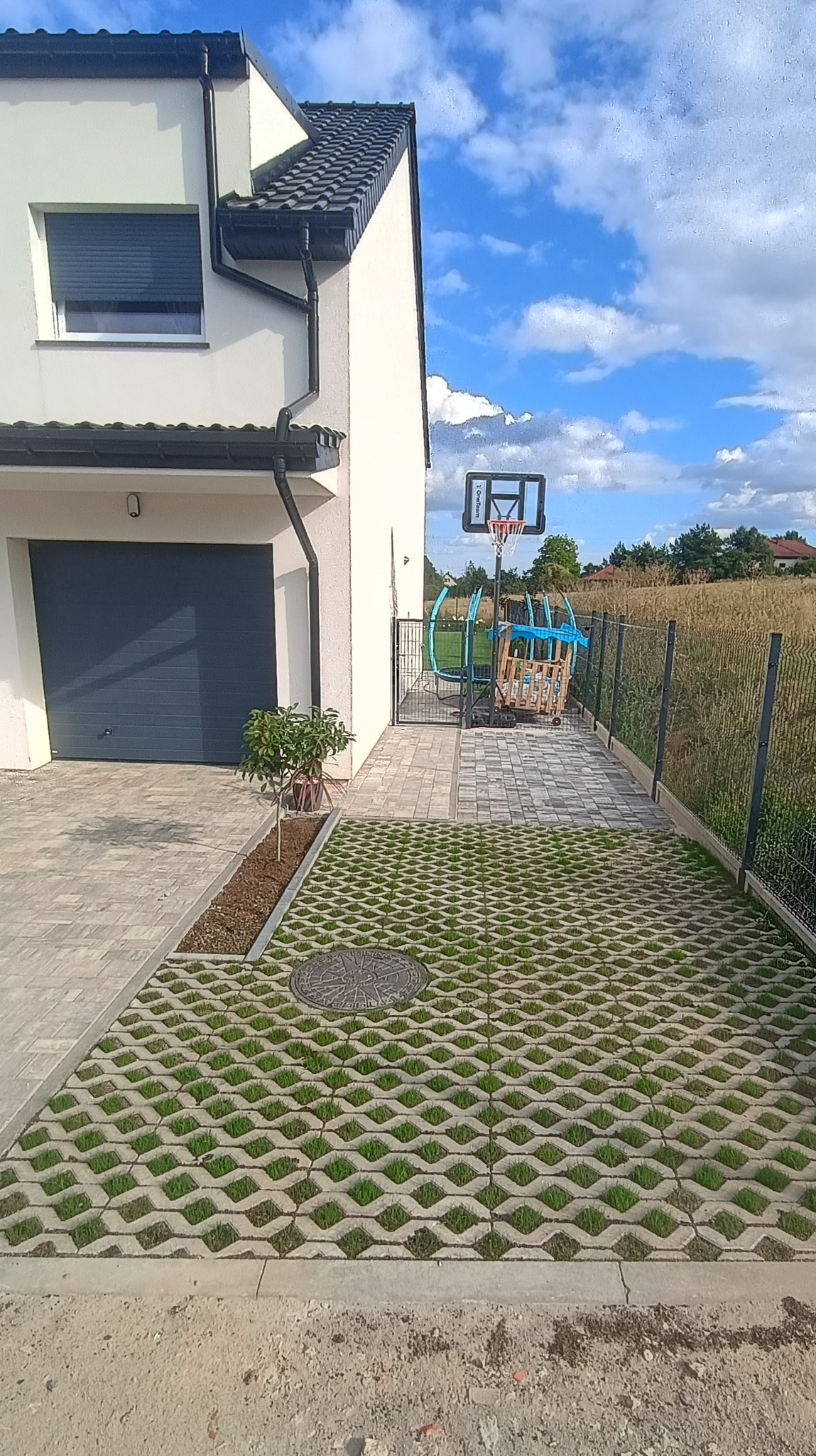 Nowoczesny podjazd z ażurowych płyt betonowych z zielenią, prowadzący do garażu i furtki z metalową siatką ogrodzeniową. W tle dom z szarą bramą garażową.