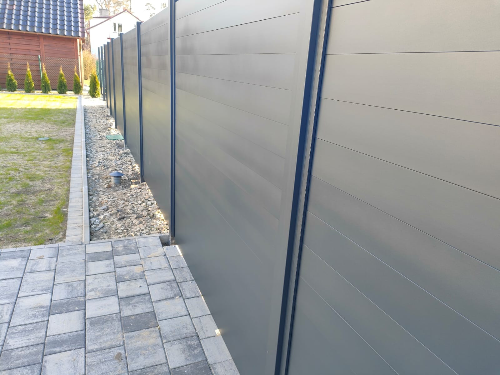 Nowoczesne ogrodzenie panelowe w kolorze szarym, z poziomymi panelami i ciemnymi słupkami, oddzielające posesję od chodnika wyłożonego kostką brukową.