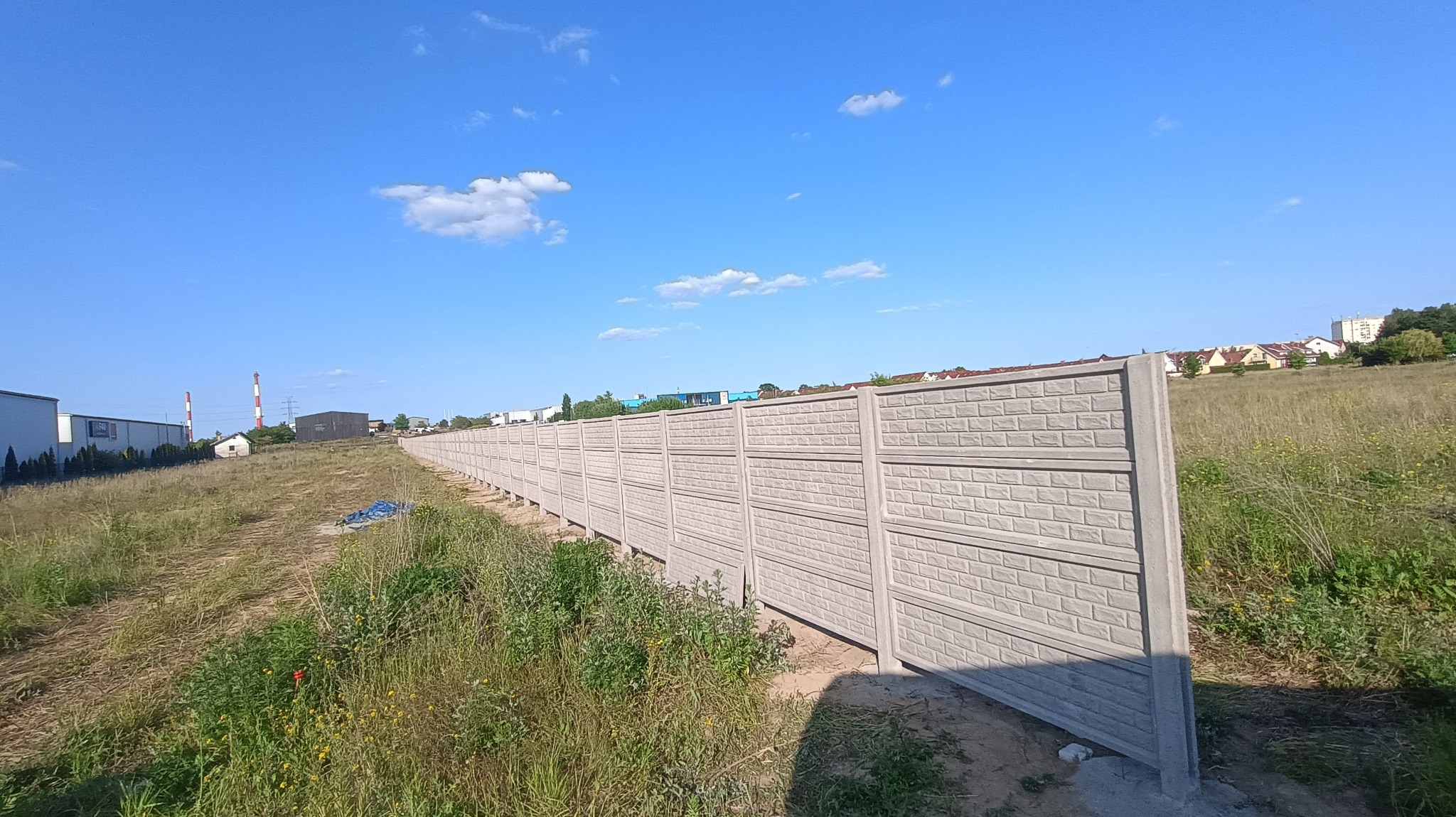 Długi, betonowy płot panelowy o fakturze cegły, oddzielający teren z trawą i chwastami od zabudowań w oddali, pod błękitnym niebem z lekkimi chmurami.