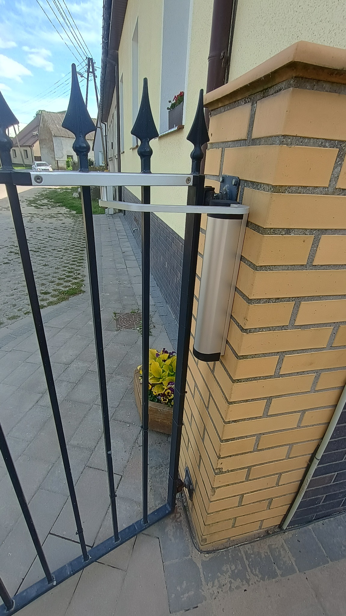 Czarna, metalowa brama z automatyką, zamontowana na ceglanym słupku. W tle widoczny fragment budynku i kostka brukowa. U dołu kadru donica z kwiatami.
