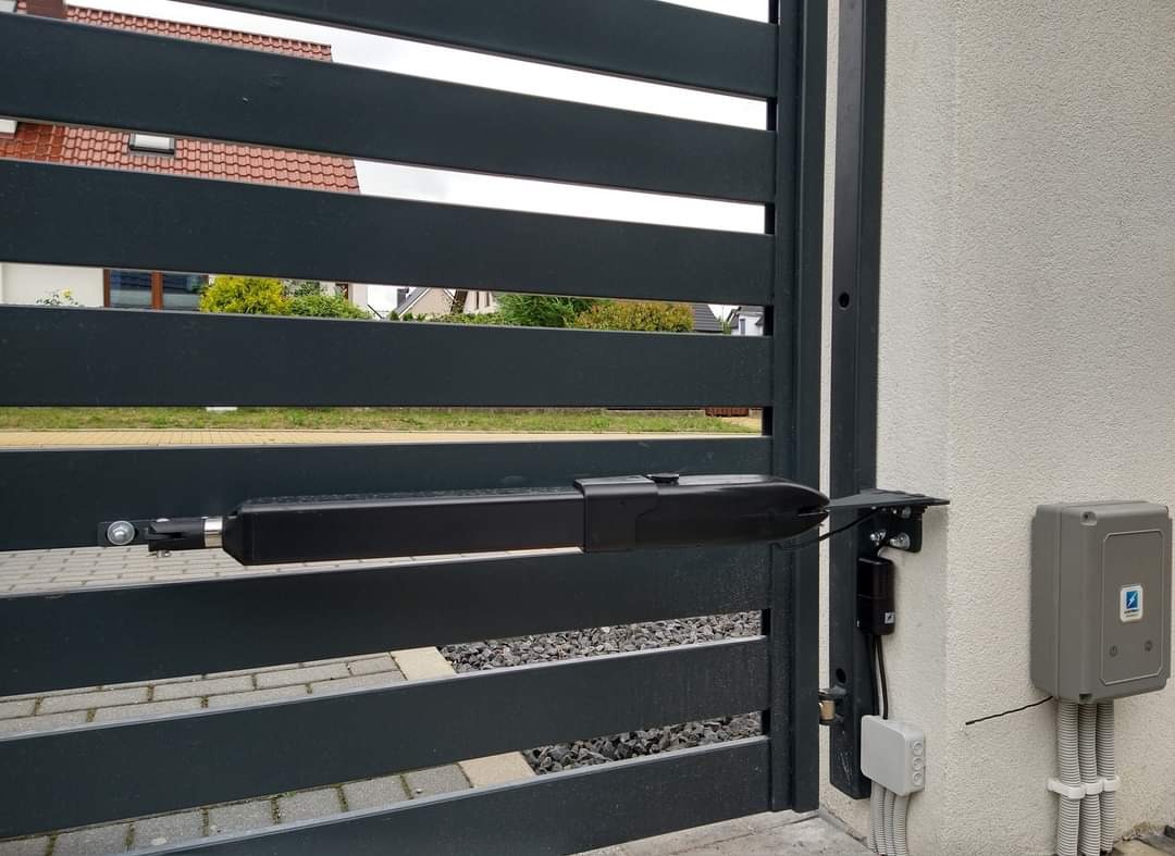 Szara, pozioma brama z automatyką, widoczny siłownik i skrzynka sterująca na tle jasnej ściany budynku. Widok z bliska na mechanizm bramy.