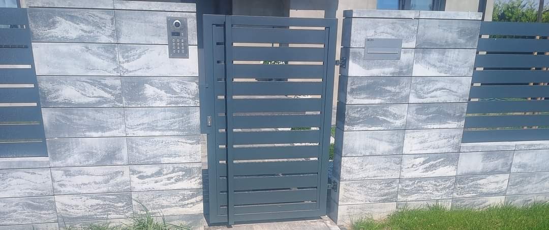 Nowoczesna, szara furtka z poziomymi szczeblami, wkomponowana w ogrodzenie z betonowych bloczków o marmurkowym wzorze. Widoczny domofon i skrzynka na listy.