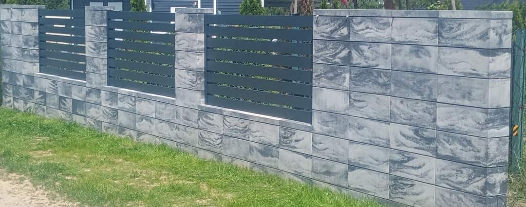 Nowoczesne ogrodzenie z szarego betonu architektonicznego i poziomych, grafitowych przęseł metalowych. Widok z poziomu gruntu, trawa w tle.