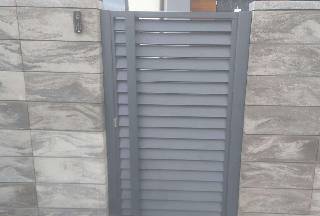 Szara, metalowa furtka lamelowa z zamkiem, wkomponowana w ogrodzenie z szarej kostki. Na słupku domofon. Nowoczesny design.