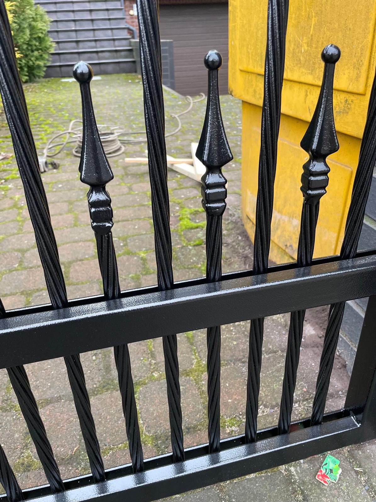 Czarny, metalowy fragment ogrodzenia z ozdobnymi grotami, widoczny z bliska na tle kostki brukowej i żółtego pojemnika. Elementy ogrodzenia skręcone.