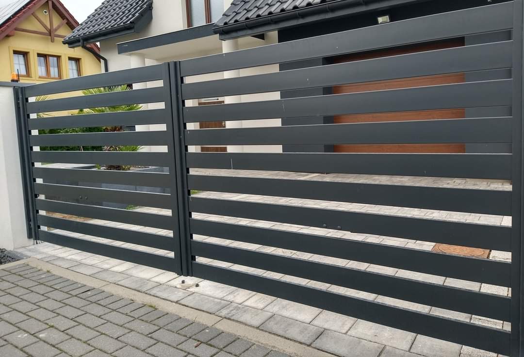 Nowoczesna, pozioma brama metalowa w kolorze grafitowym, z widocznymi elementami mocującymi do kostki brukowej. W tle fragment domu z jasną elewacją i ciemnym dachem.