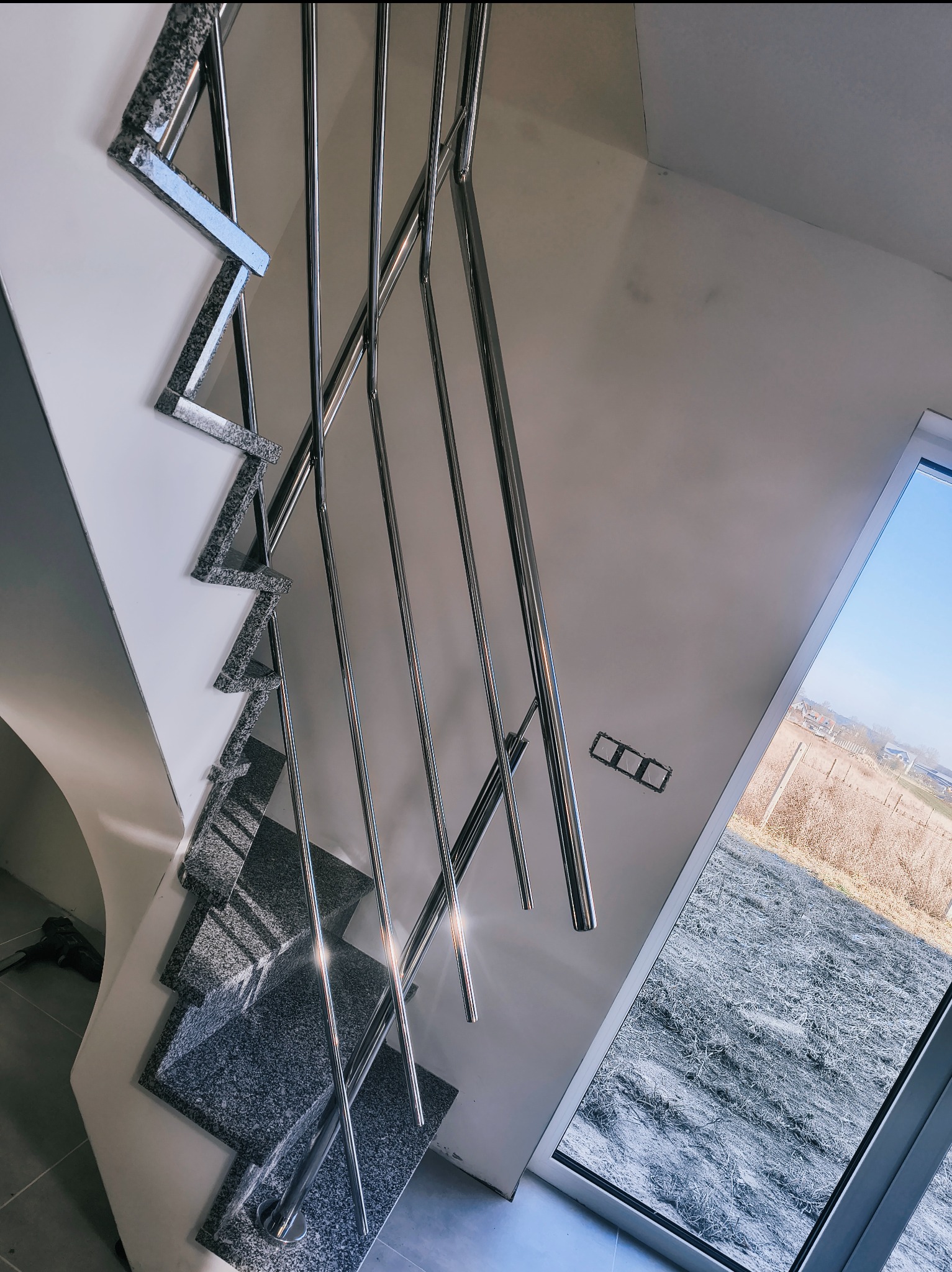 Nowoczesne schody z balustradą ze stali nierdzewnej, wykończone granitem. Widok z dołu, z oknem w tle. Polerowane elementy odbijają światło.