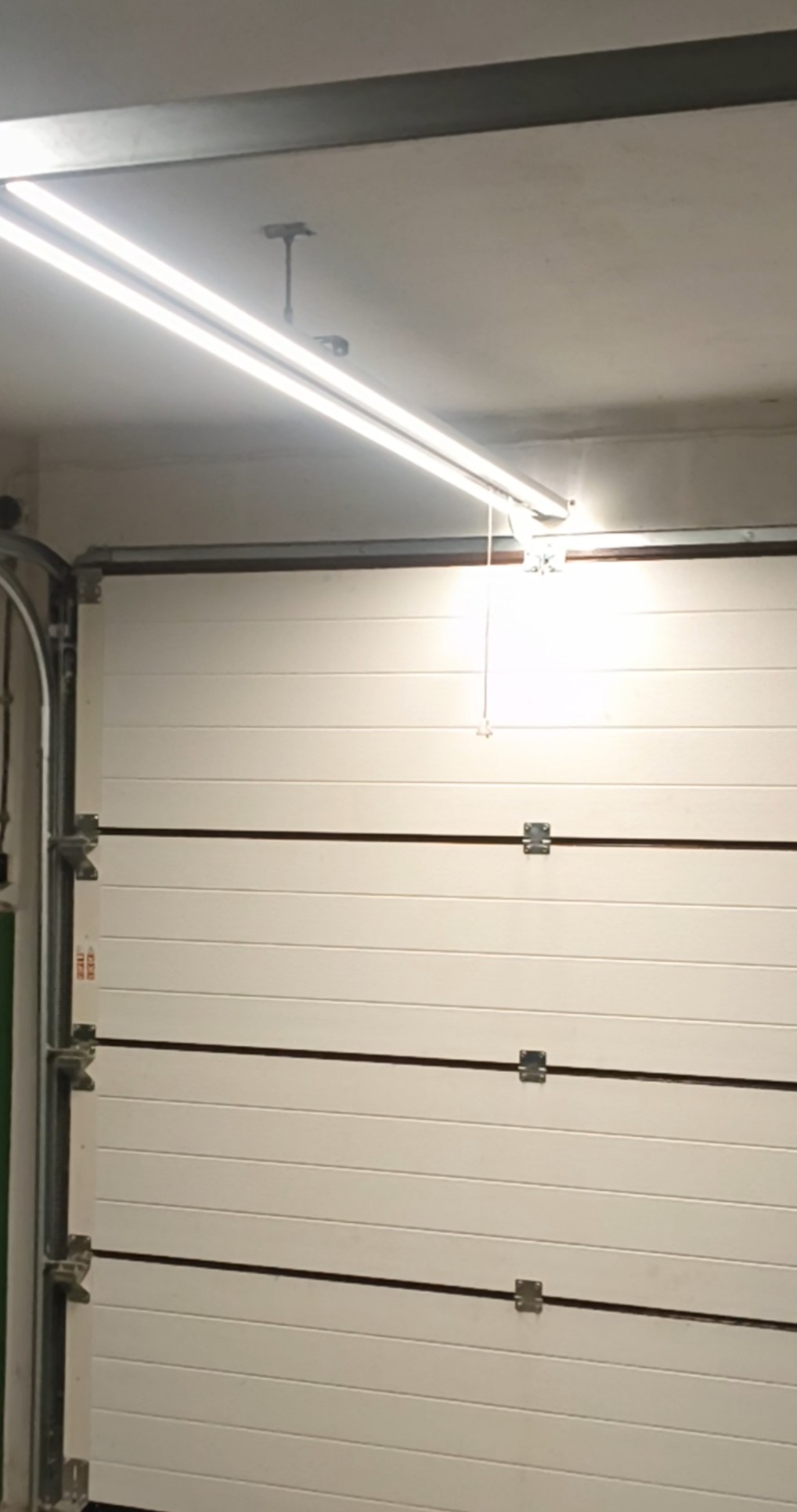 Otwarta, segmentowa brama garażowa w kolorze białym, oświetlona podwójną lampą LED. Widoczne metalowe prowadnice i mocowania. Brama w pozycji otwartej, widok od wewnątrz garażu.