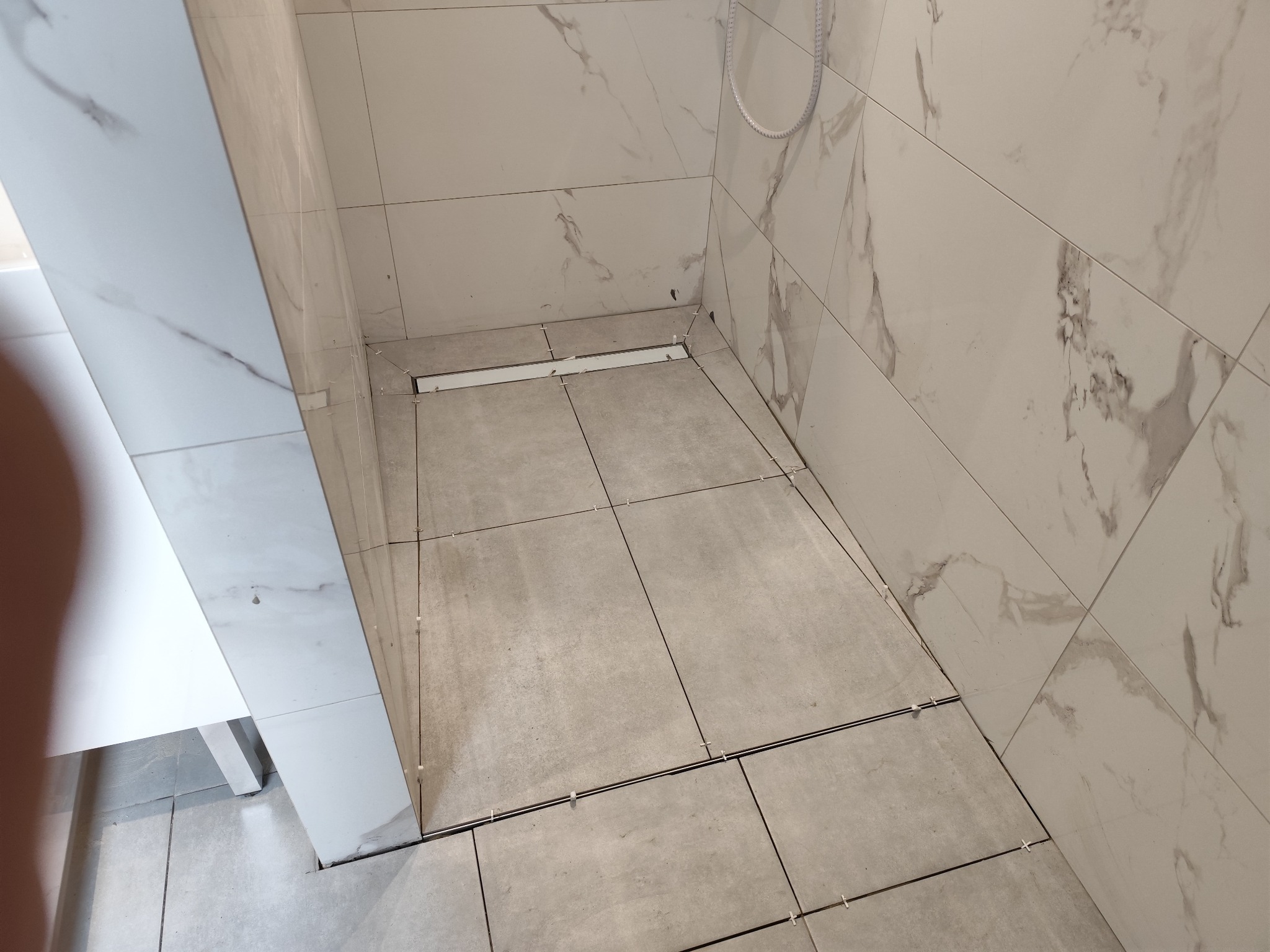 Ułożone płytki podłogowe w łazience z odpływem liniowym, widoczne krzyżyki dystansowe. Ściany wyłożone białymi płytkami imitującymi marmur.