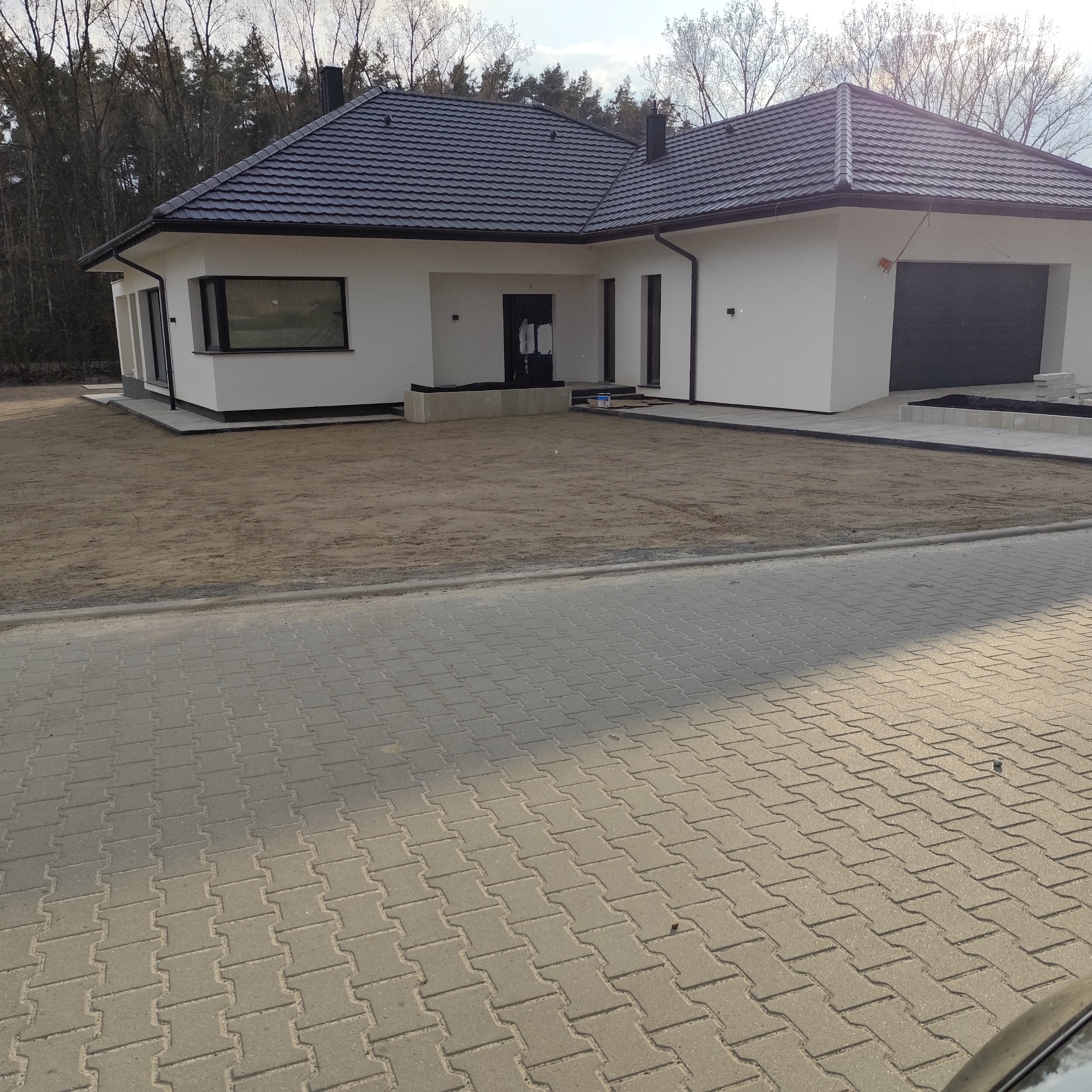 Nowoczesny dom jednorodzinny z czarnym dachem i białymi ścianami w Sulechowie. Widoczny fragment chodnika i przygotowany teren pod ogród.