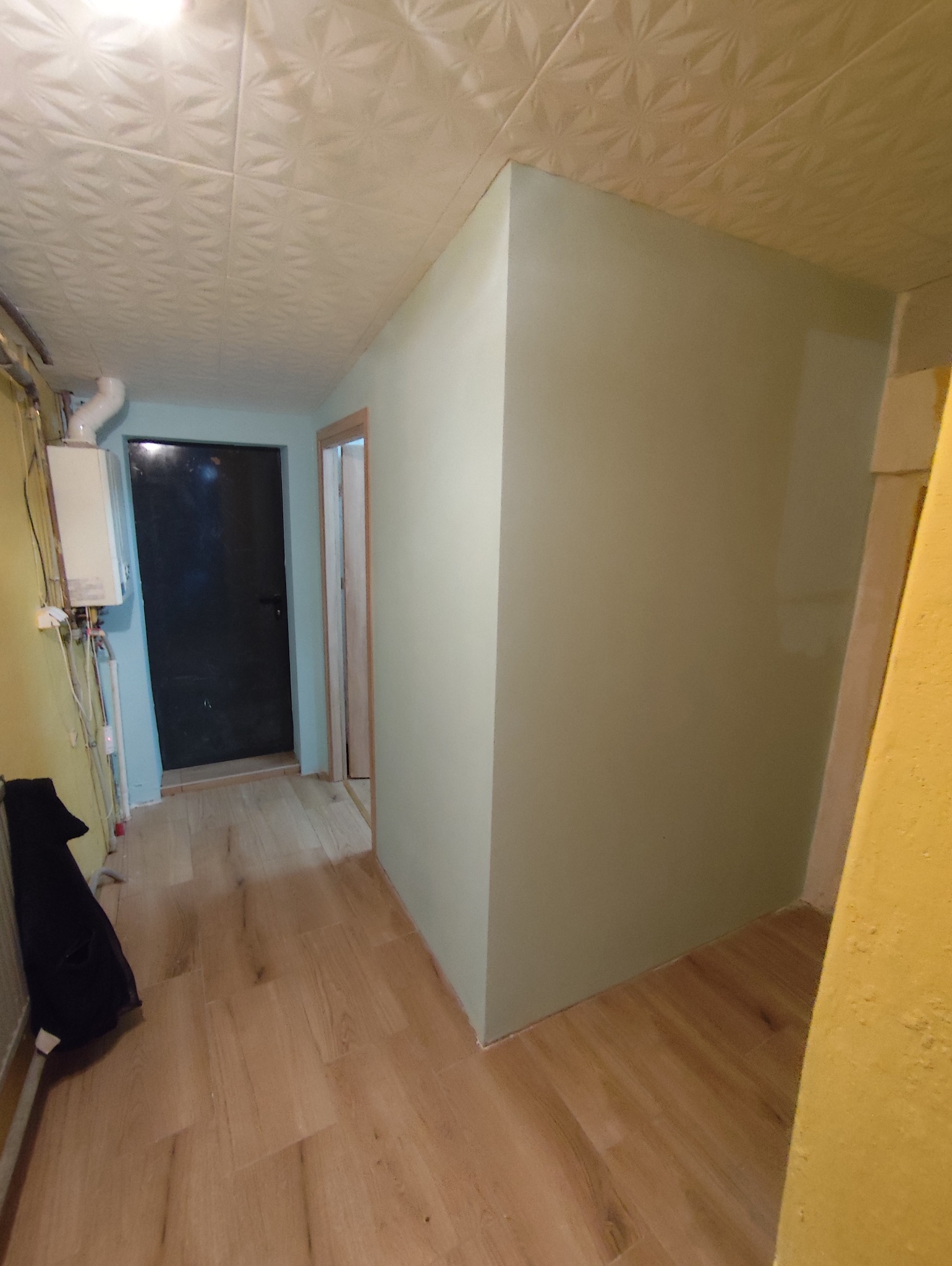 Korytarz po malowaniu ścian na jasny odcień zieleni, z nową podłogą imitującą drewno. Widoczny fragment grzejnika i drzwi wejściowe.