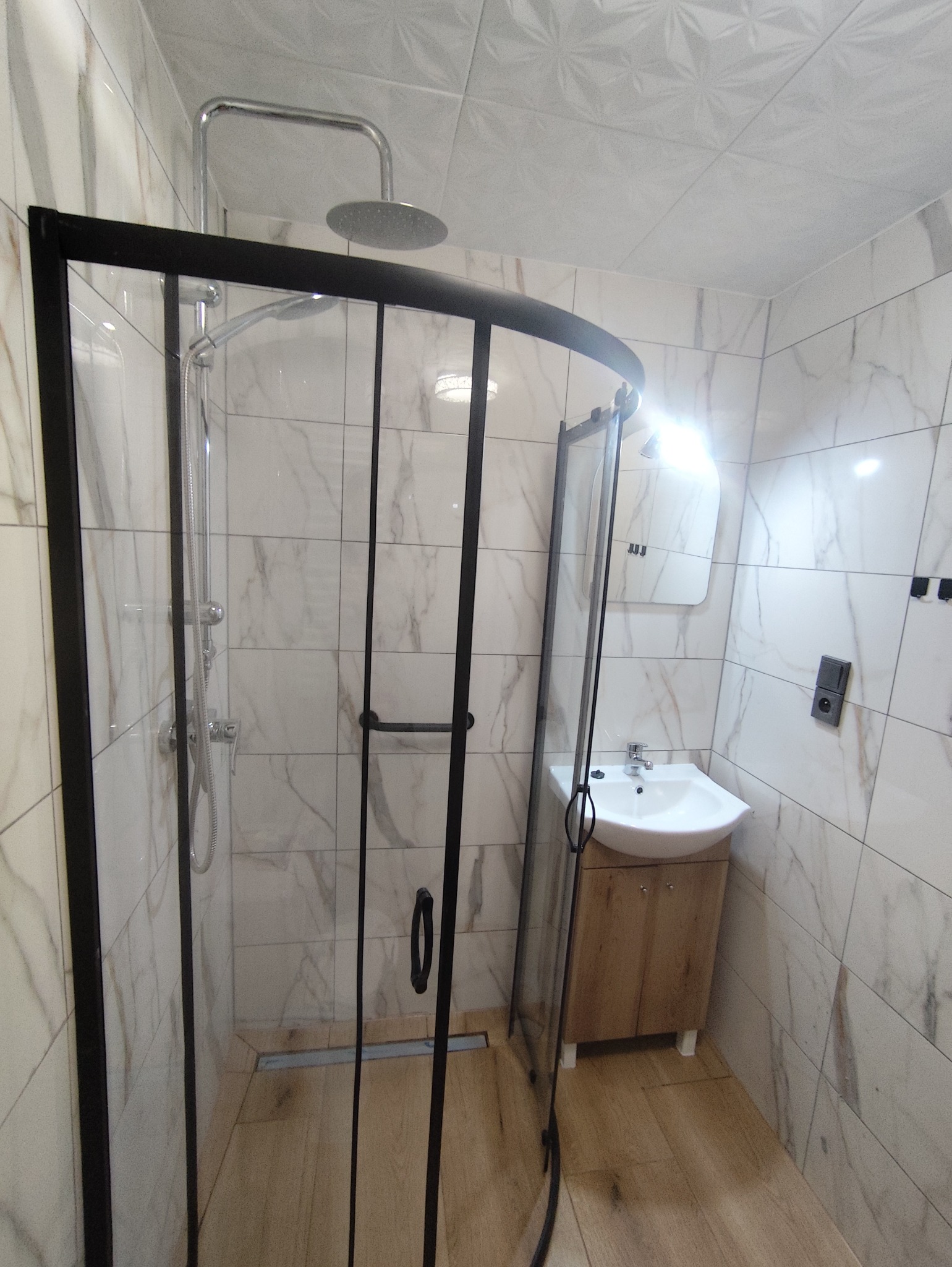 Wykończona łazienka z kabiną prysznicową typu walk-in z czarnymi profilami, umywalką z szafką w odcieniach drewna i marmurowymi płytkami na ścianach.