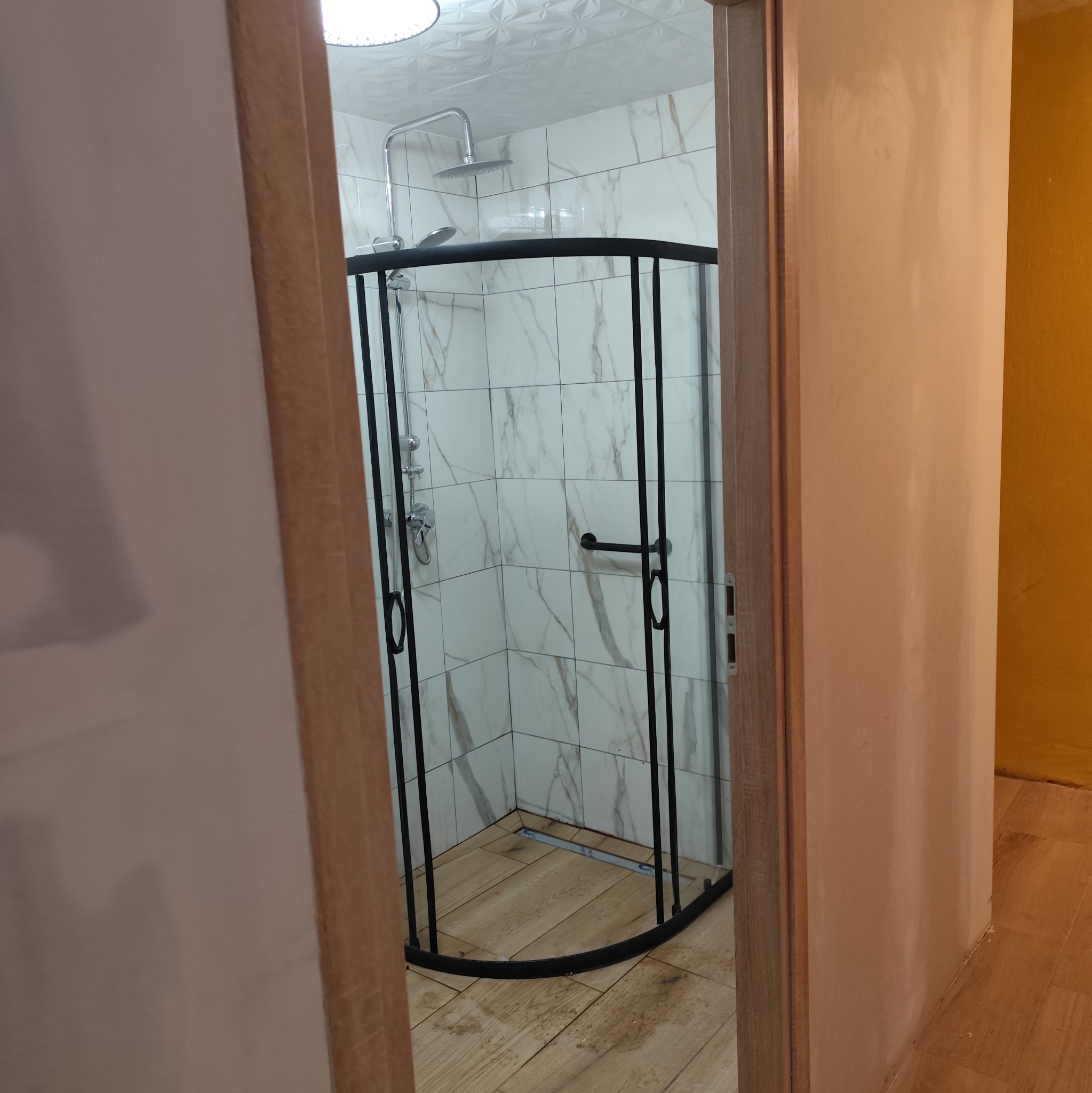 Nowoczesna kabina prysznicowa z czarną ramą i marmurowymi płytkami w wykończonej łazience, widoczna zza otwartych drzwi. Jasne drewno na podłodze.