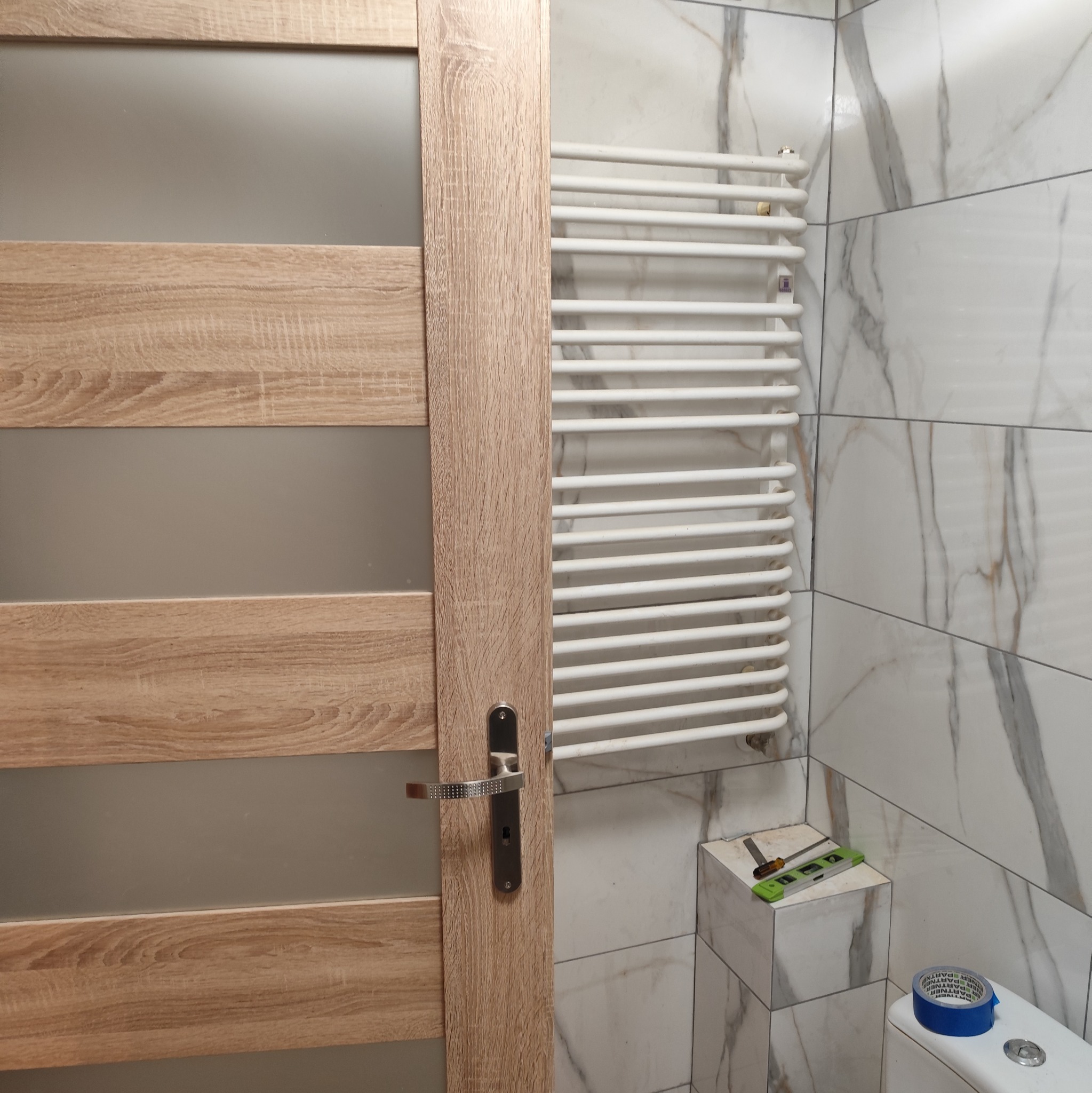 Wykończona łazienka z grzejnikiem drabinkowym na marmurowych płytkach, widoczna część drzwi z drewnianą okleiną i narzędzia glazurnicze na półce.