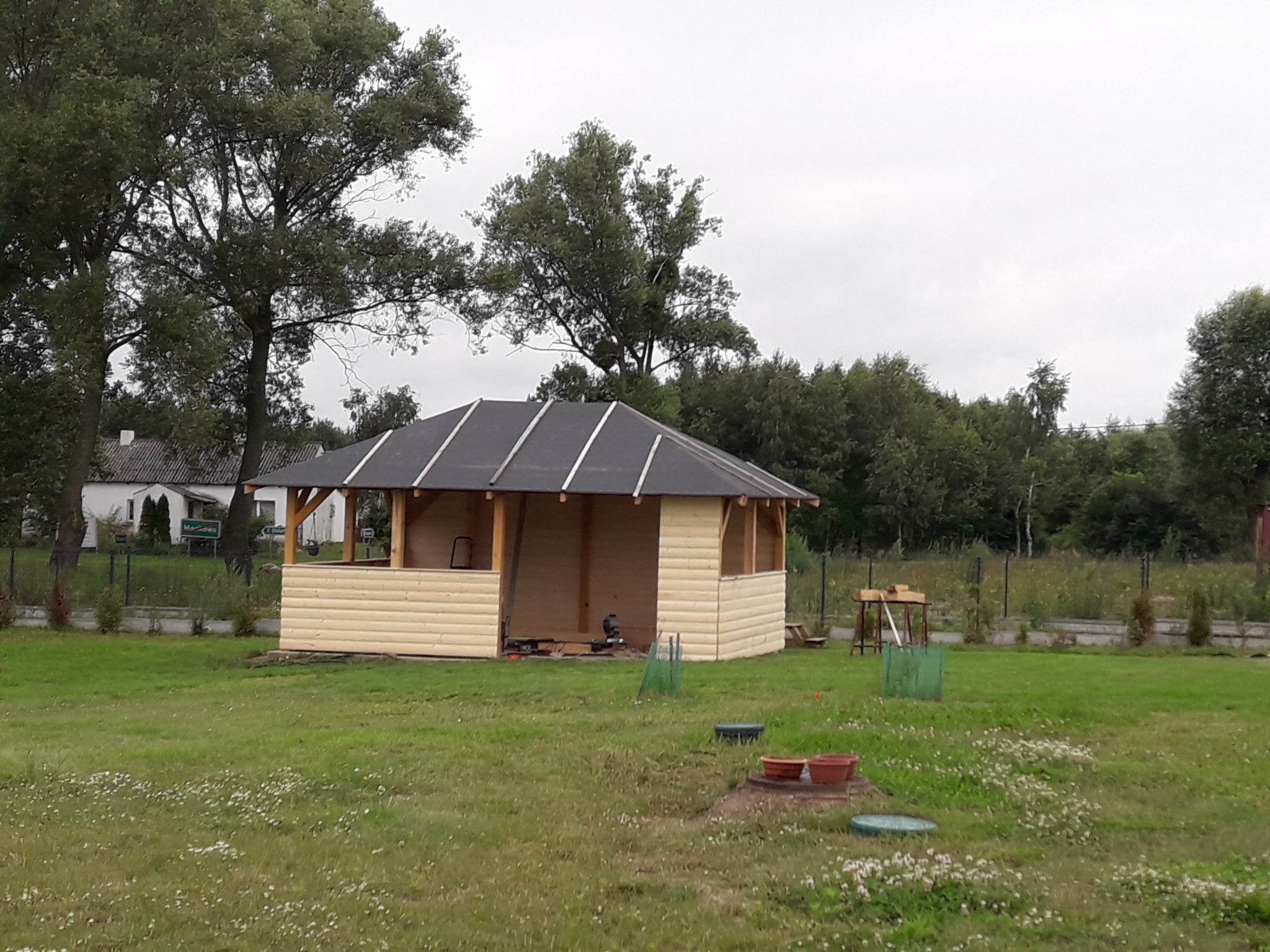 Drewniana altana z czarnym dachem w trakcie budowy na zielonej trawie, narzędzia widoczne wewnątrz. W tle drzewa i dom.