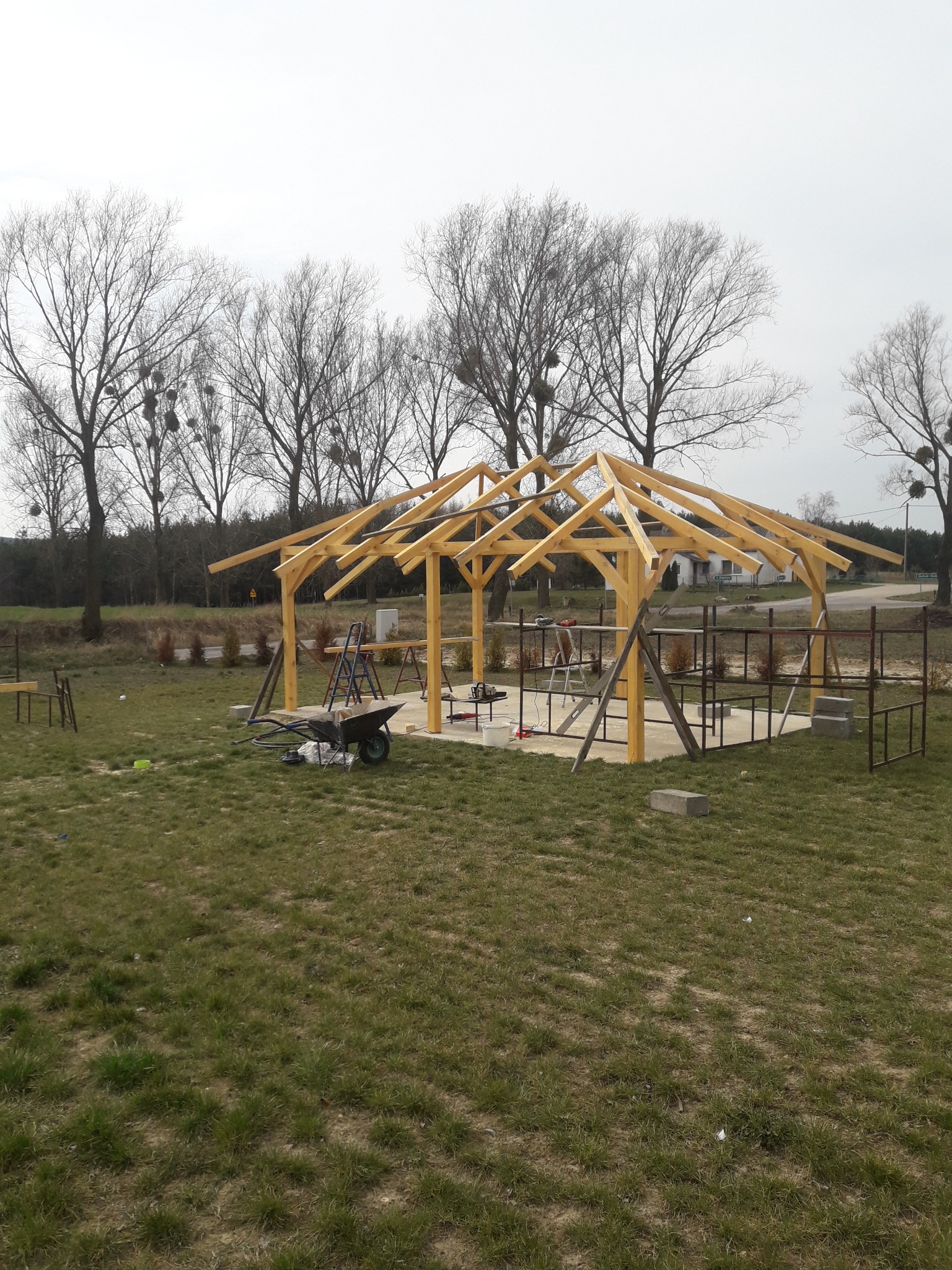 Konstrukcja drewnianej altany w trakcie budowy na trawniku, widoczne narzędzia i drabiny. W tle drzewa bez liści i budynek mieszkalny.