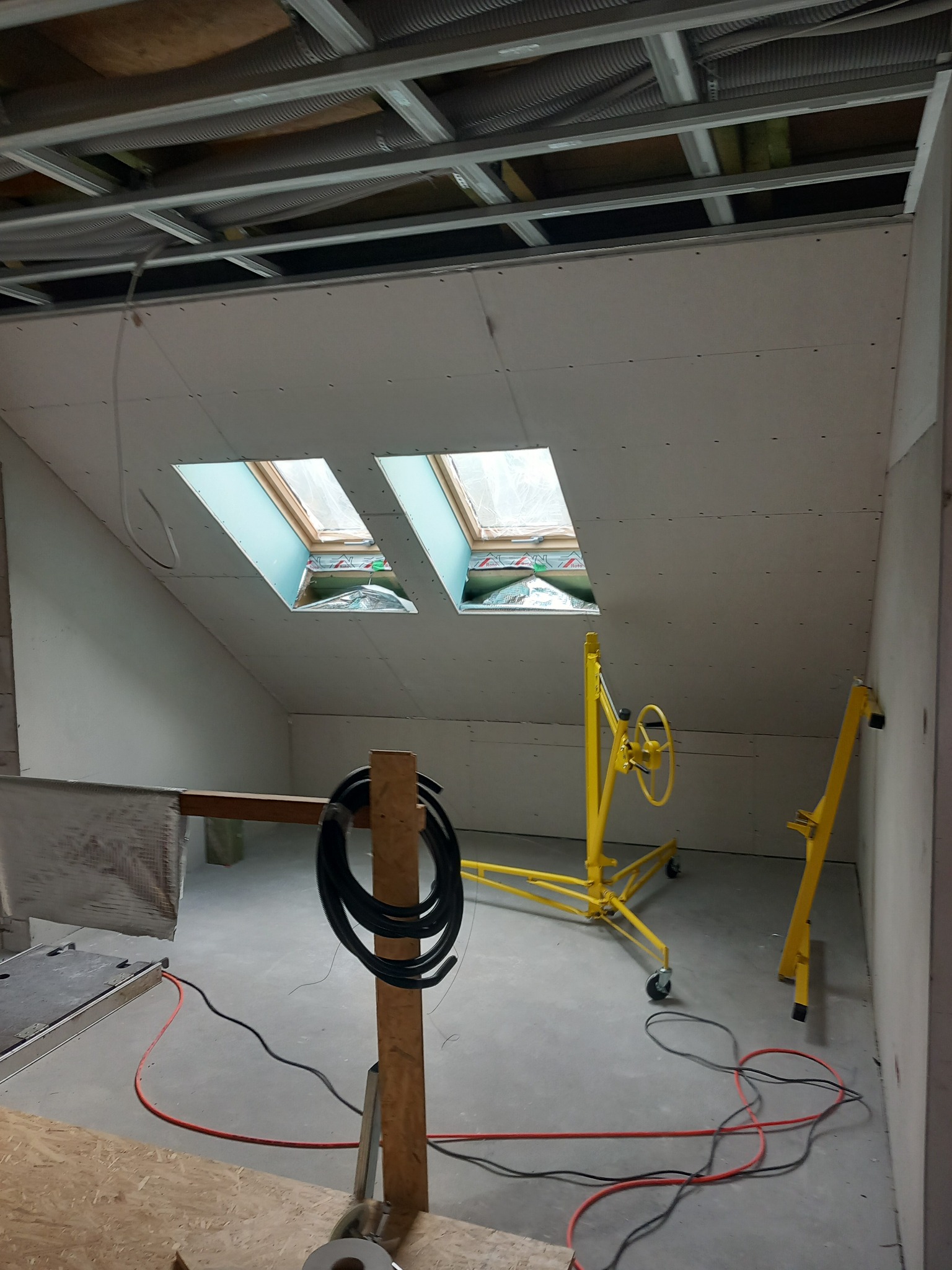 Poddasze w trakcie wykańczania: zamontowane płyty gipsowe, widoczne okna dachowe, metalowa konstrukcja sufitu i żółty podnośnik do płyt. Przewody elektryczne na podłodze.