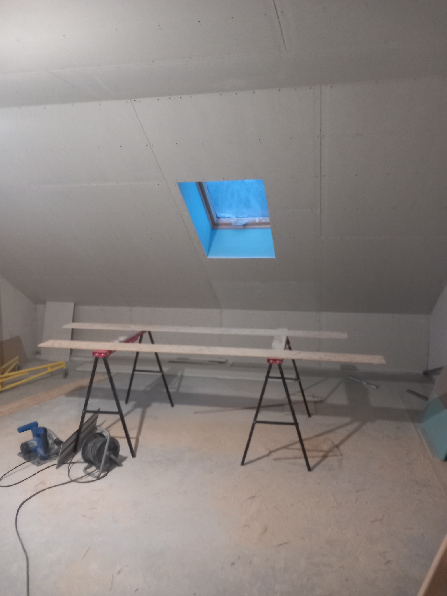 Poddasze w trakcie wykańczania: płyty gipsowe na skosach, okno dachowe z niebieską folią, narzędzia i deski na kozłach. Widok na surowy stan przed dalszymi pracami.