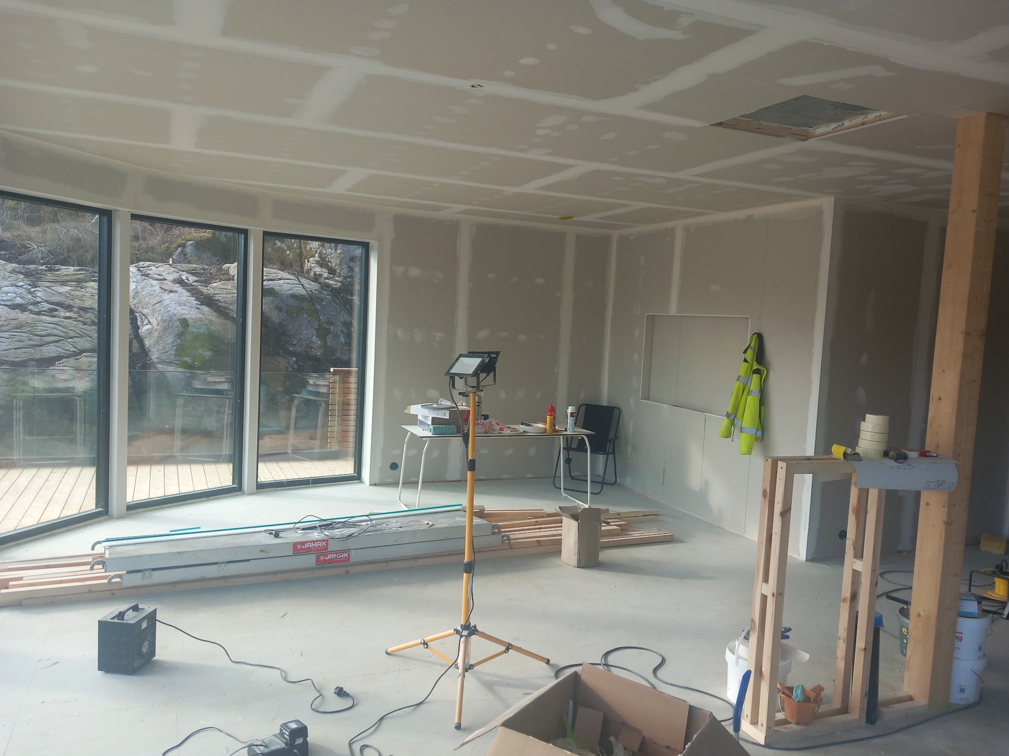 Wnętrze w trakcie wykończenia: płyty gipsowe na ścianach i suficie, widoczne narzędzia i materiały budowlane, duże okna z widokiem na skały. Etap szpachlowania.