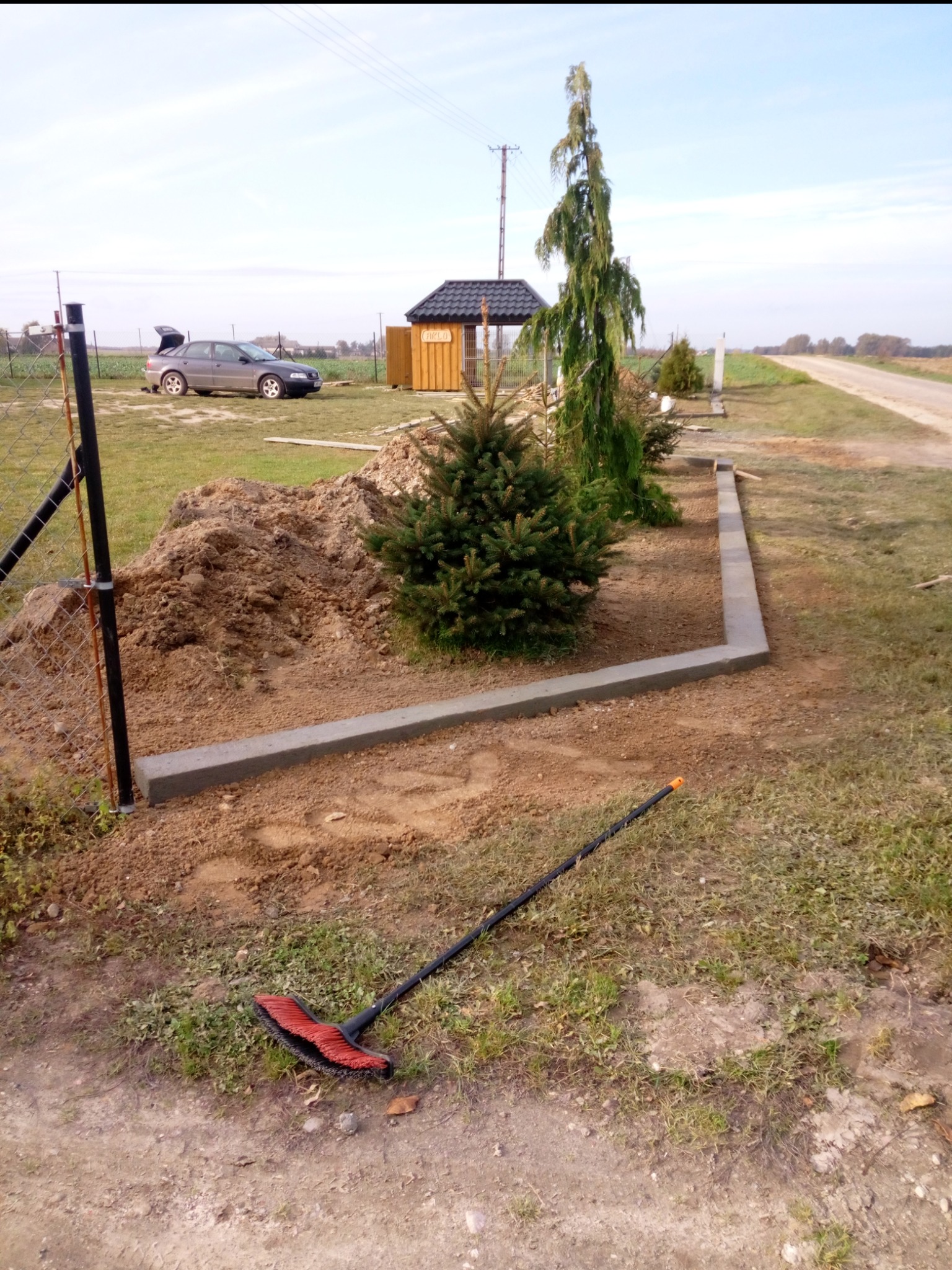 Układanie obrzeży trawnikowych z betonu, widoczne młode drzewka iglaste, w tle samochód osobowy i drewniana altana. Prace ziemne w toku, widoczna miotła.