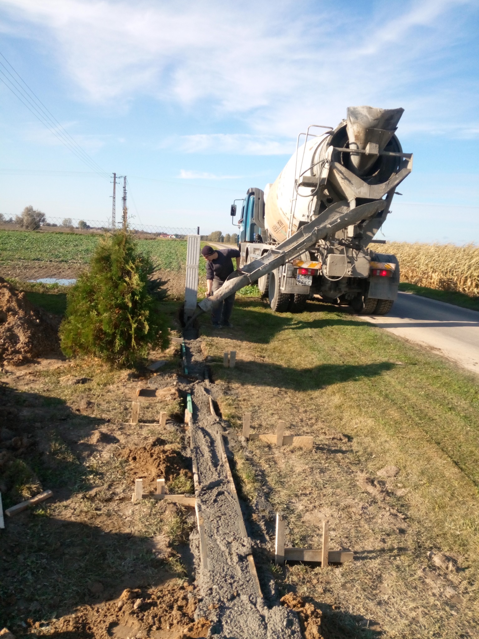 Wylewanie betonu z betoniarki do wąskiego wykopu z drewnianym szalunkiem na zewnątrz. Prace ziemne w słoneczny dzień. Widoczny pracownik i maszyna.