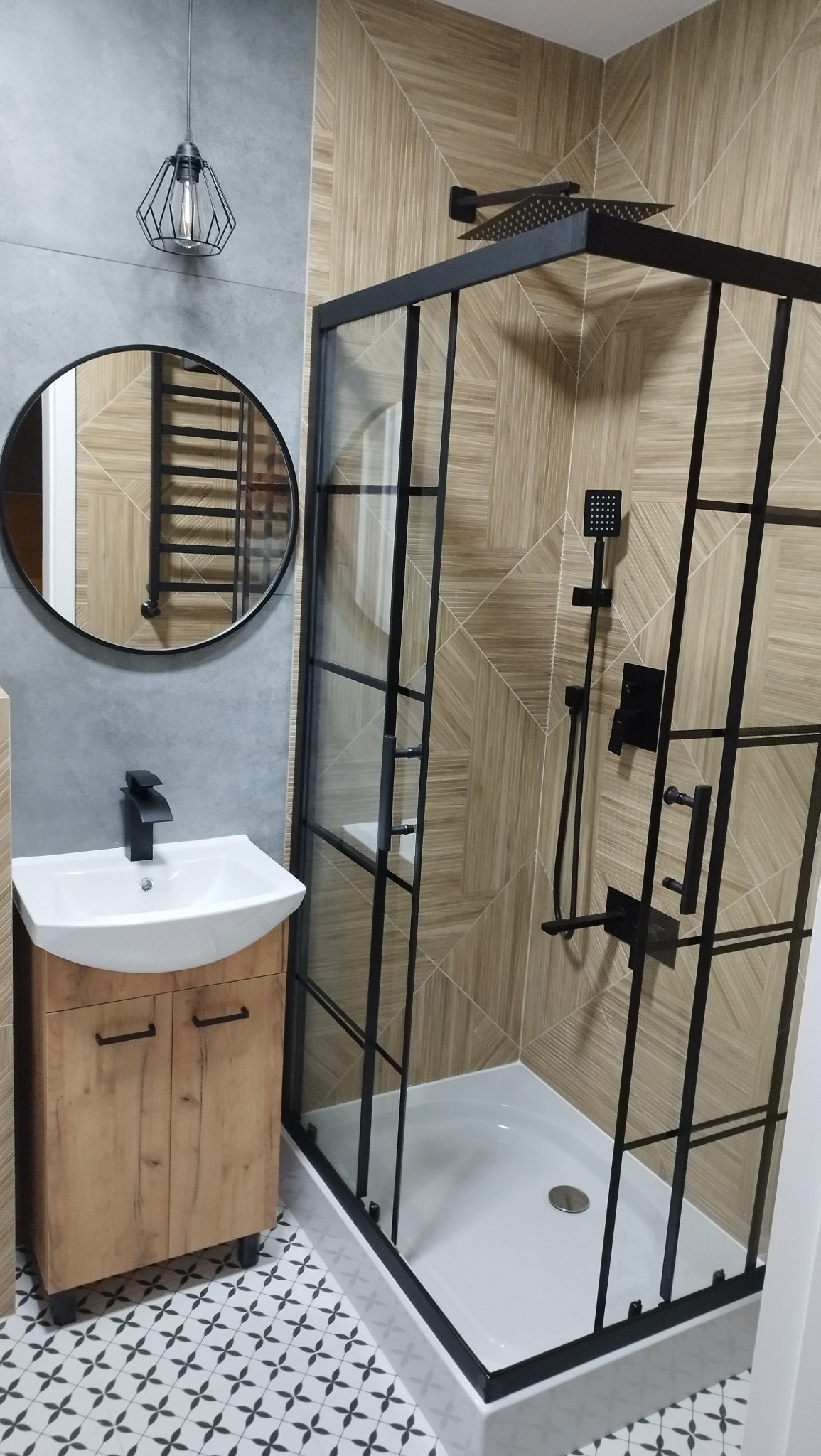 Nowoczesna łazienka z kabiną prysznicową typu walk-in z czarnymi profilami, umywalką z szafką w kolorze drewna i okrągłym lustrem na szarej ścianie.