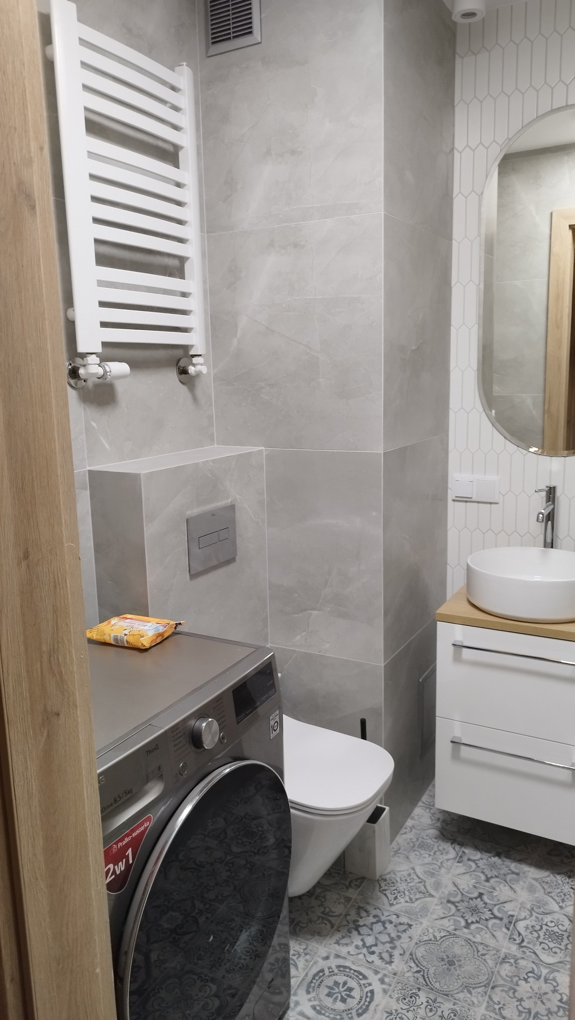 Wykończona łazienka w szarościach: płytki imitujące marmur, owalne lustro, umywalka nablatowa, grzejnik drabinkowy i pralka z wzorem na drzwiczkach.