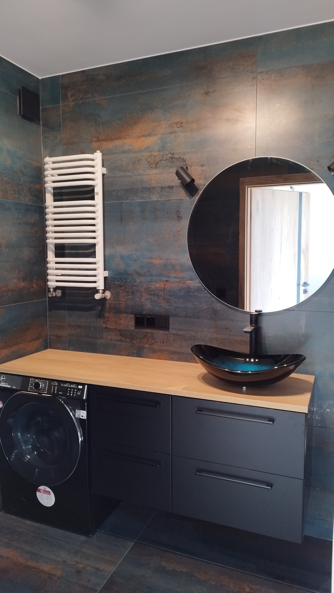Nowoczesna łazienka z umywalką w kształcie misy, szafką z drewnianym blatem i pralką. Na ścianie płytki imitujące rdzę i okrągłe lustro.