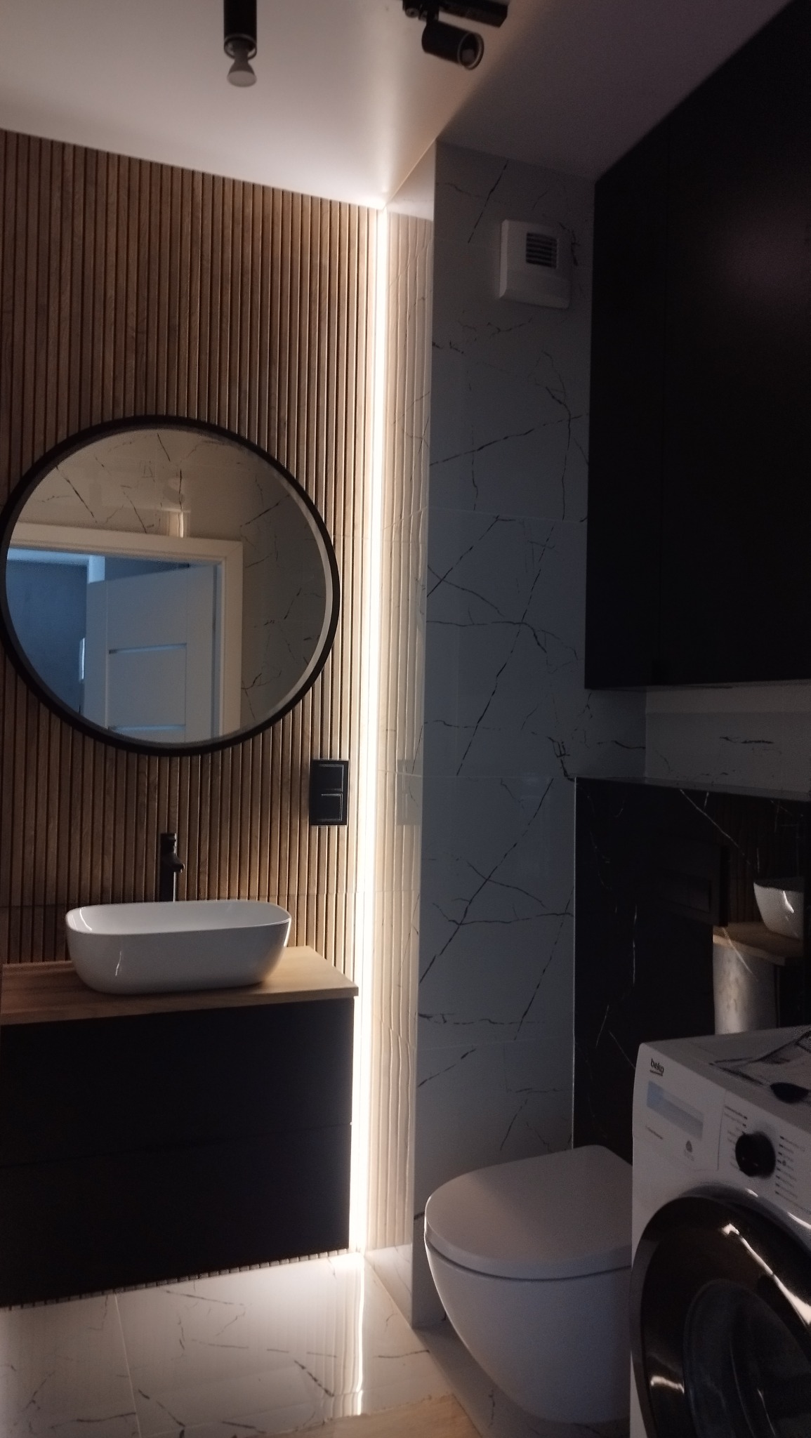 Nowoczesna łazienka z drewnianą ścianą, owalnym lustrem i podświetleniem LED. Umywalka nablatowa i marmurowe płytki tworzą elegancki wystrój.
