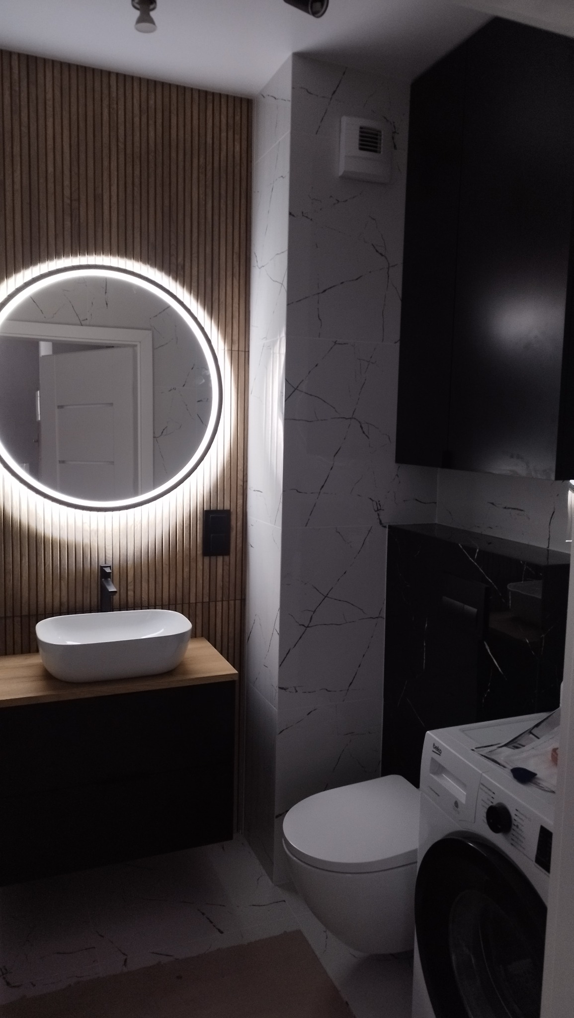 Nowoczesna łazienka z okrągłym lustrem LED, drewnianą ścianą i marmurowymi płytkami. Umywalka nablatowa i czarne szafki dodają elegancji.