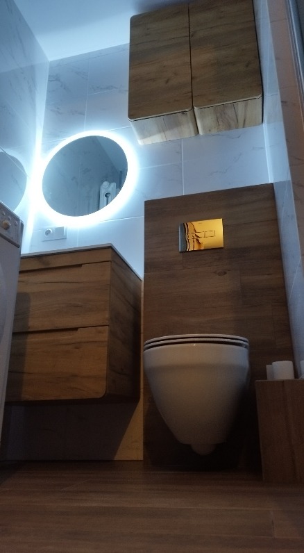 Nowoczesna łazienka z podwieszaną toaletą, szafką pod umywalką i okrągłym lustrem z podświetleniem LED. Płytki imitujące drewno na podłodze i ścianie.