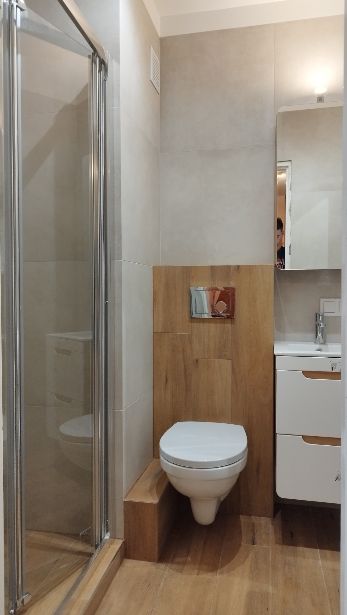 Nowoczesna łazienka po remoncie: kabina prysznicowa ze szklanymi drzwiami, podwieszana toaleta z drewnianym panelem, biała szafka z umywalką i lustro.