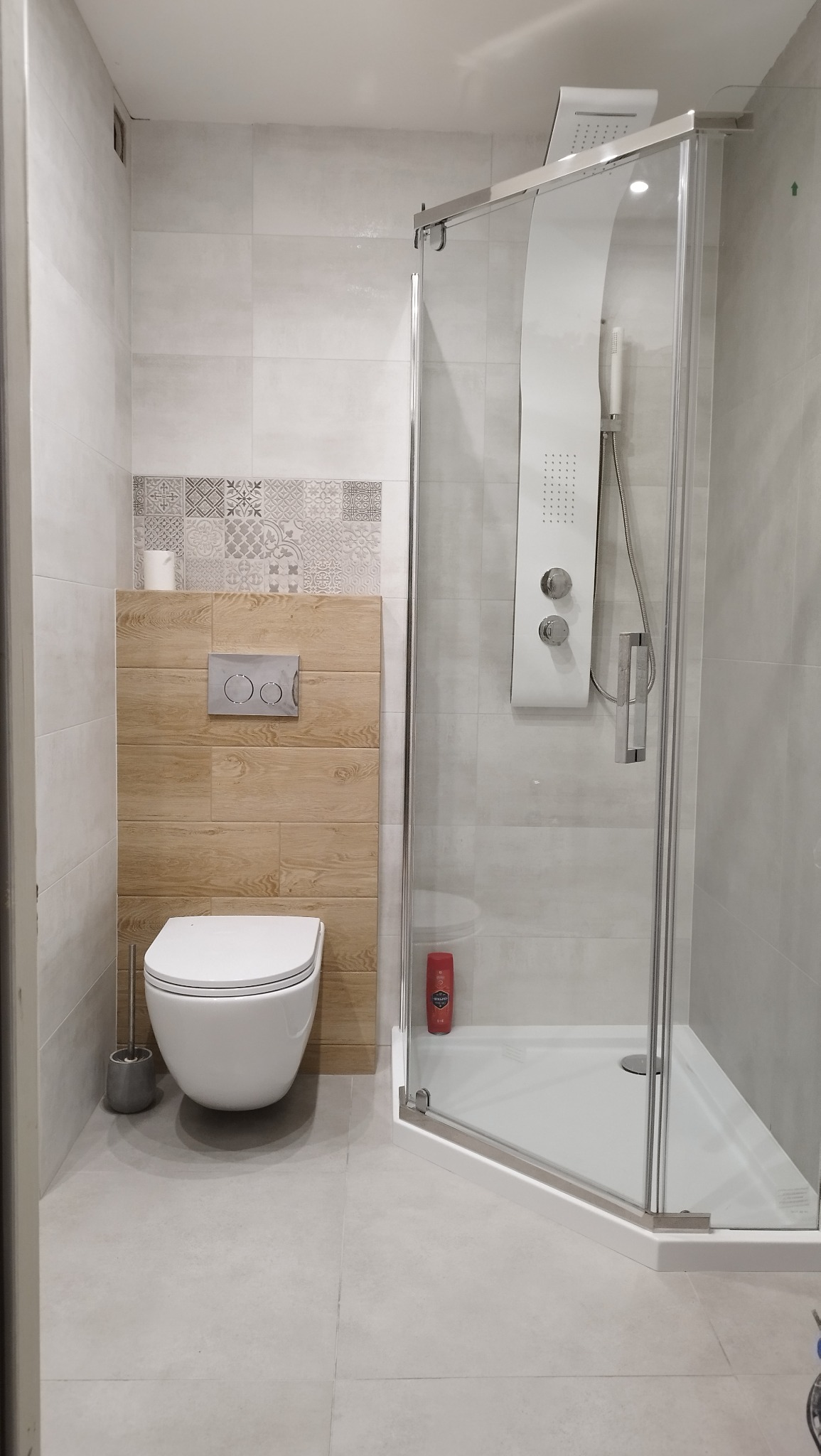 Nowoczesna łazienka po remoncie: kabina prysznicowa z hydromasażem, podwieszana toaleta z drewnianą obudową i dekoracyjnymi płytkami. Jasne płytki podłogowe i ścienne.