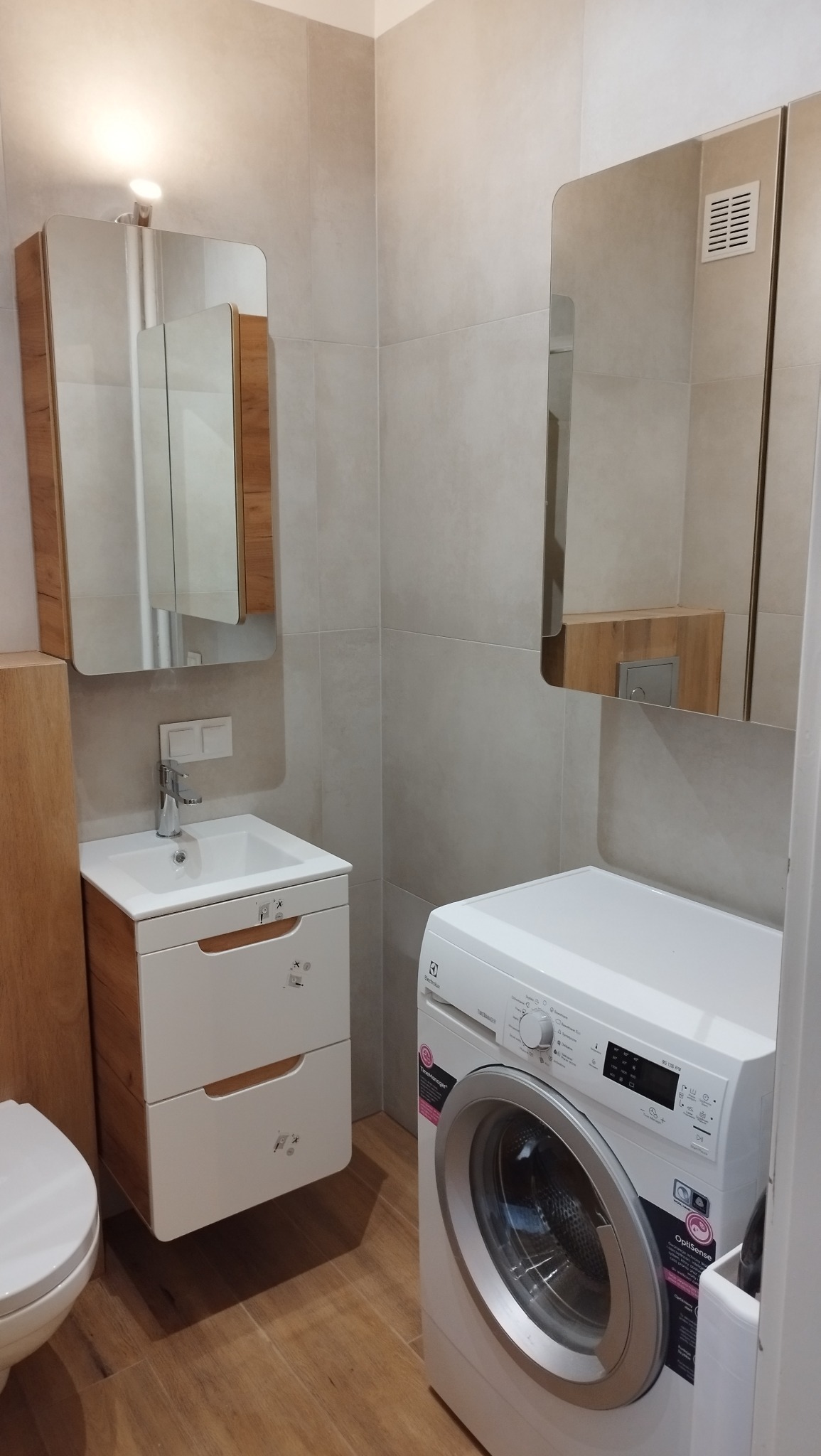 Jasna łazienka z drewnianymi akcentami: szafka pod umywalką, lustro z oświetleniem, pralka Electrolux i beżowe płytki podłogowe.