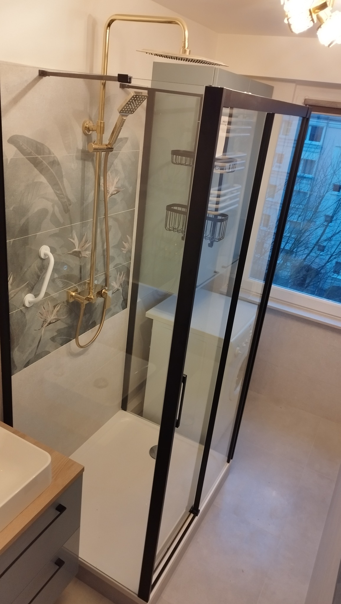 Nowoczesna kabina prysznicowa z czarnymi profilami i złotą armaturą na tle płytek z motywem roślinnym. Widoczny brodzik i umywalka z szafką.