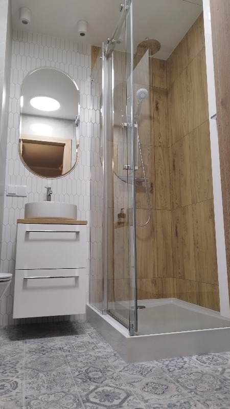 Nowoczesna łazienka z kabiną prysznicową typu walk-in, umywalką nablatową na szafce i lustrem owalnym. Biało-drewniana kolorystyka, wzorzysta podłoga.