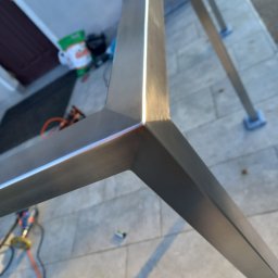 Metalsystem Barbara Brzezińska - Fragment konstrukcji metalowej, prawdopodobnie pergoli, z widocznymi spawami i elementami montażowymi na tle kostki brukowej i narzędzi.